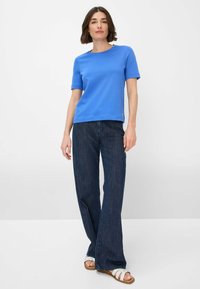 T-shirt blu a maniche corte in cotone misto, abbinato a pantaloni larghi in denim scuro e sandali bianchi con cinturini. Design semplice, senza fantasie.