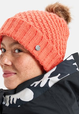 Nahaufnahme einer lächelnden Person, die eine orange gestrickte Mütze mit Bommel und einen schwarz-weiß gemusterten Jackenkragen trägt.