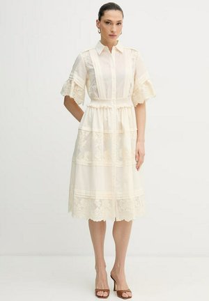 Donna con un abito midi in pizzo color crema, maniche corte, chiusura frontale con bottoni, vita aderente e tacchi aperti marroni, in piedi su sfondo bianco.