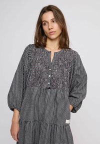 Barna hajú nő, aki hosszú ujjú, fekete-fehér kockás ruhát visel, melynek a mellrésze ráncolt és gombokkal díszített, sima háttér előtt áll.