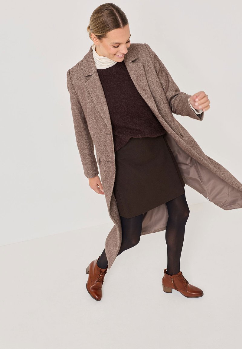 Manteau en laine mélangée marron, à coupe longue et avec revers, superposé sur un pull sombre et une jupe, associé à des bottines marron.