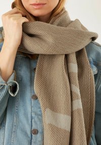 Écharpe tissée beige avec un motif texturé, présentant des accents plus clairs. Tissu doux drapé lâchement autour du cou, associé à une veste en denim.