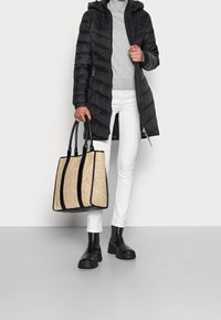 Persona che indossa un cappotto trapuntato nero, un maglione grigio, pantaloni bianchi, stivali neri, tenendo una grande borsa tote beige con manici e bordi neri.
