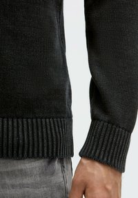 Nahaufnahme einer Hand und eines Handgelenks einer Person, die einen schwarzen, gerippten Strickpullover und hellgraue, strukturierte Hosen trägt.