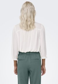 Leichte, weiße Bluse mit einem gerafften Rückendetail und 3/4-Ärmeln. Kombiniert mit grünen Hosen, die einen elastischen Bund und Gesäßtaschen haben.