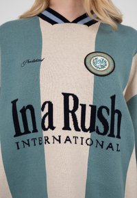 Femeie purtând un pulover cu dungi în culoarea teal și bej, cu textul „In a Rush International” și un patch rotund pe piept.