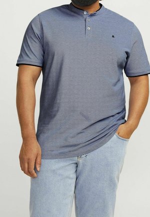 Homme portant un t-shirt henley bleu-gris à manches courtes avec deux boutons, associé à un jean bleu clair, une main dans la poche.