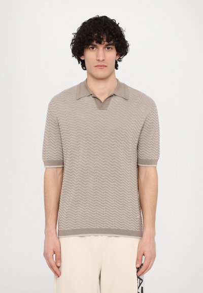 Beige neulepolo, jossa on sahalaitakuviota, lyhyet hihat ja V-pääntie. Korostaa teksturoitua pintaa sekä erottuvia resoreita hihoissa ja helmassa.