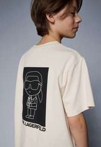 Beige kokvilnas T-krekls ar melnu grafiku, uz kuras attēlota stilizēta figūra saulesbrillēs aizmugurē, ar tekstu "KARL LAGERFELD".