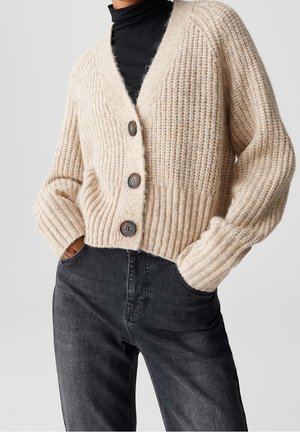 Personne portant un cardigan beige en grosse maille avec trois gros boutons sur un col roulé noir et un jean gris foncé, les mains dans les poches.