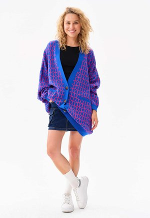 Kobieta uśmiechająca się, ubrana w oversize'owy kardigan z niebiesko-różowym wzorem, czarny top, dżinsowa spódnica, białe skarpetki i białe trampki, stojąca ze skrzyżowanymi nogami.