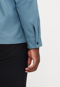 Una camicia azzurra chiara con polsi abbottonati e una fessura laterale. Il tessuto ha una consistenza liscia, accentuata da un design aderente.