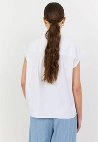 Blusa bianca a maniche corte con taglio dritto e tessuto morbido, con inserto sul retro e spacchi laterali, abbinata a pantaloni azzurri chiari.