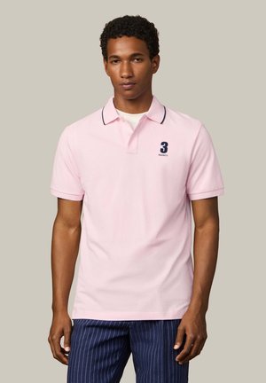 Man draagt een lichtroze poloshirt met een marineblauw nummer 3 en tekst op de borst, gecombineerd met marineblauwe broek met krijtstrepen.