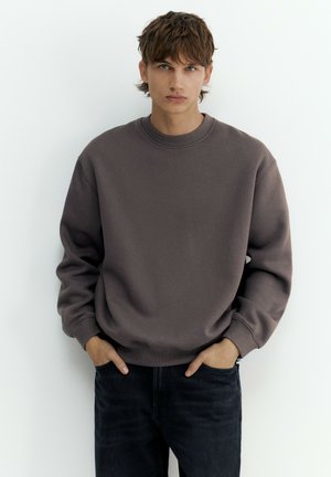Junger Mann mit braunen Haaren, der einen braunen übergroßen Pullover und schwarze Jeans trägt, mit den Händen in den Taschen, vor weißem Hintergrund.