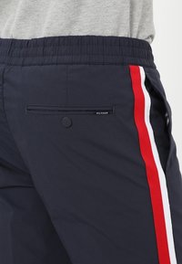 Marinblå atletiska byxor med röda och vita sidostråkar, elastisk midja, bakficka med knappstängning och diskret logodetalj.