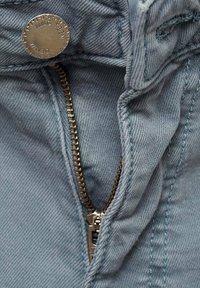 Close-up van lichtblauwe spijkerbroek met gedeeltelijk open koperen rits en metalen knoop met het label "DENIM NMG TEEN."