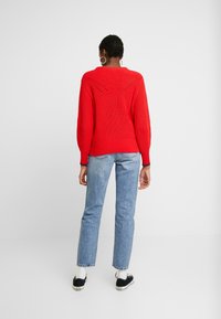 Rød strikket trøje med ribbet tekstur, dyb V-formet design bagpå, kombineret med lyseblå jeans med lige ben og sorte sneakers.