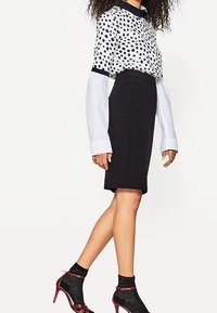 Zwarte kokerrok gecombineerd met een witte blouse met zwarte stippen en lange mouwen. Geaccentueerd met rode hoge hakken en gemusterde sokken.
