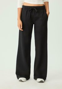 Schwarze weit geschnittene Hose mit Kordelzugbund, Seitentaschen und glatter Textur, kombiniert mit weißen Sportschuhen.