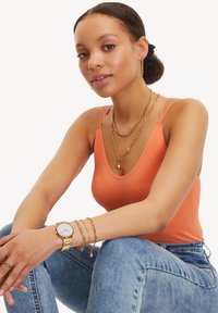 Oranges, geripptes Tanktop, übereinander getragene goldene Halsketten und eine Uhr mit weißem Ziffernblatt. Getragen mit blauen Denim-Jeans. Glatte Textur, taillierte Passform.