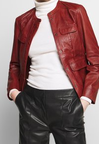 Röd läderjacka med rund hals, tryckknappsstängning och två framfickor, parat med en vit ribbad polotröja och svarta läderbyxor.