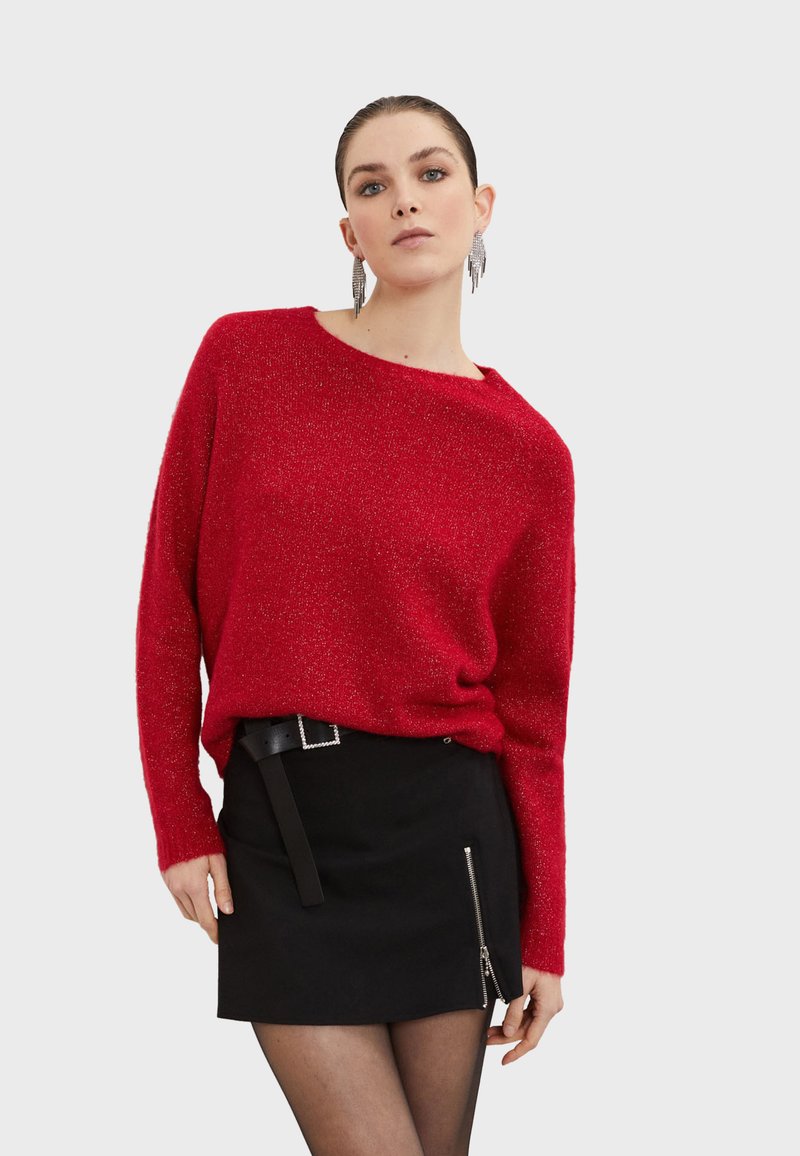 Roter Strukturpullover mit langen Ärmeln und weitem Ausschnitt, kombiniert mit einem schwarzen Minirock mit seitlichem Reißverschluss und einem Gürtel.