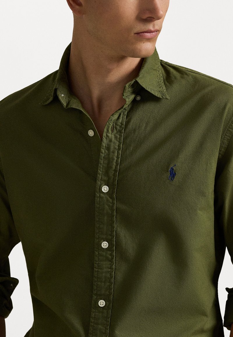 Olive green button-up shirt made of textured cotton, featuring a small blue logo, buttoned collar, and a slim-fit design.
