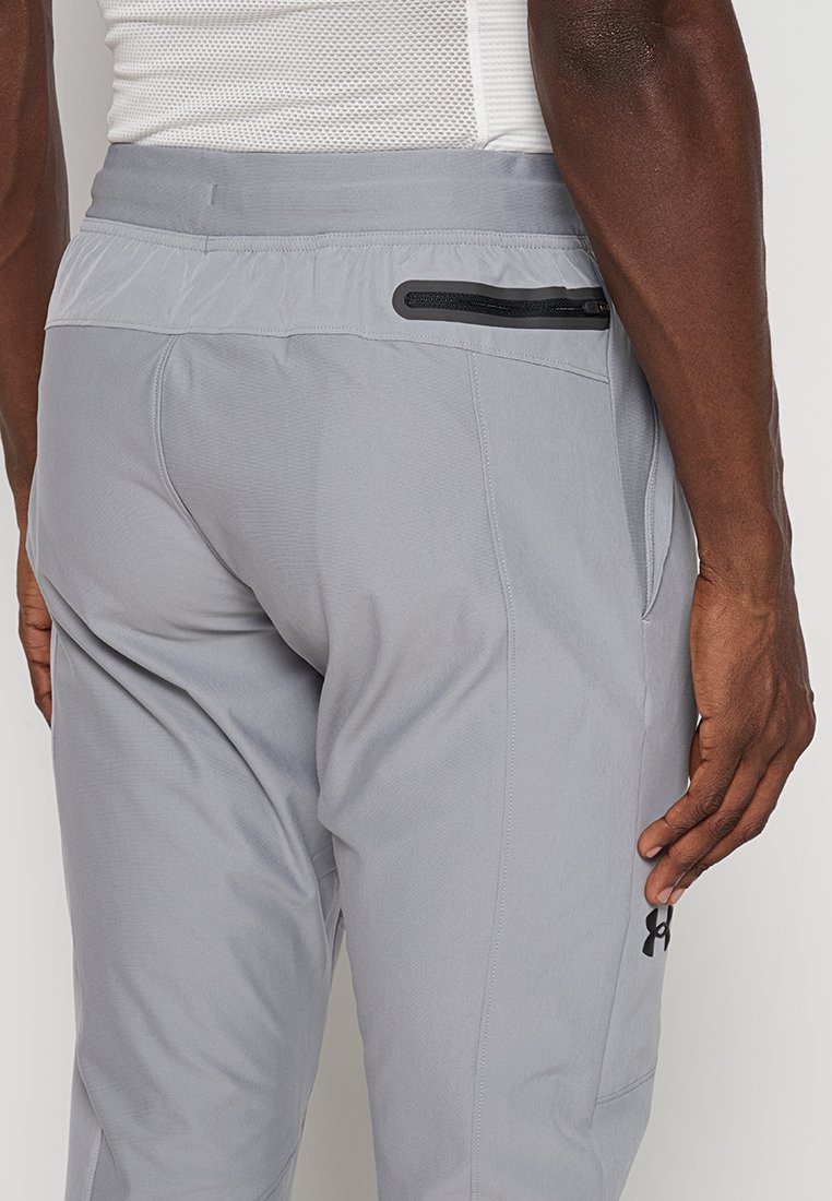 Un pantalon de sport gris en tissu léger présente une taille avec un cordon de serrage caché, une poche arrière zippée et des coutures latérales.