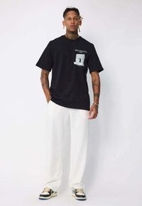 Homme tatoué portant un t-shirt graphique noir de la Renaissance, un pantalon blanc et des baskets noir et blanc, se tenant devant un fond blanc.