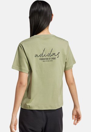 Person mit langen dunklen Haaren, die ein hellgrünes Adidas T-Shirt mit der Aufschrift „adidas FOUNDATION OF SPORT“ auf dem Rücken trägt, kombiniert mit schwarzen Hosen.