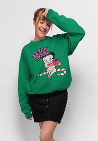Sweatshirt oversize vert avec un graphisme de dessin animé de Betty Boop, arborant des couleurs vives et un texte audacieux au-dessus du personnage. Jupe en denim noir.