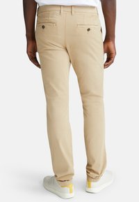Pantaloni beige in misto cotone con vestibilità slim, dotati di due tasche posteriori con chiusura a bottone e polsini affusolati. Abbinati a sneakers bianche.