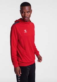 Rød hætte sweatshirt fremstillet af tekstureret stof, med et logo på brystet og sorte detaljer på skuldrene. Båret med sorte bukser.