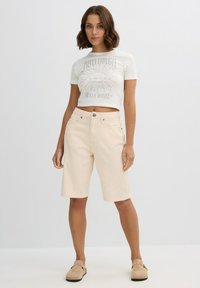 Shorts de mezclilla beige claro con un ajuste relajado, que cuentan con un cierre de botón y bolsillos. Combinados con una camiseta gráfica recortada de color blanco roto.