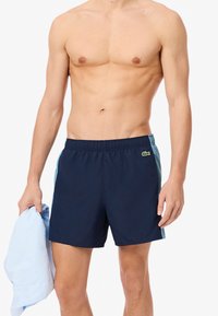 Marineblauwe zwempantalons met een elastische tailleband, zijaccenten in het blauw en een klein logo op de voorkant, gecombineerd met een lichtblauw shirt.