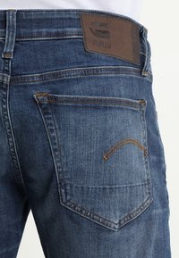 Dark blue denim jeans with a textured finish. Features include contrast stitching, a brown leather label, and back pockets with curved stitching.