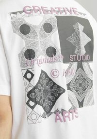 Camiseta de algodón blanca con diseños gráficos abstractos en negro y gris, y texto rosa que dice “CREATIVE STUDIO ORIGINALS™ © 1990.”