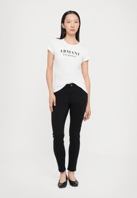 Balts īsām piedurknēm T-krekls ar "Armani Exchange" logo, apvienots ar pieguļošām melnām džinsēm un melniem balerīnu apaviem, uz vienkārša balta fona.