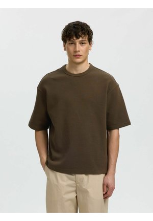 Jeune homme aux cheveux courts et bouclés, portant une chemise marron texturée à manches courtes et un pantalon beige, debout avec une main dans la poche.