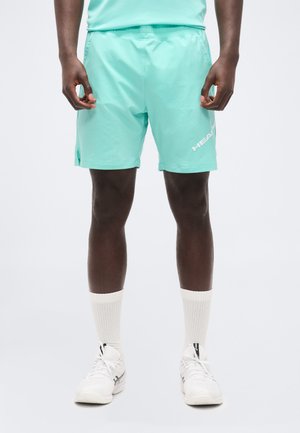 Moški, oblečen v svetlo turkizne športne kratke hlače z belim logotipom, bele nogavice in bele športne copate, stoji pred enobarvnim ozadjem.