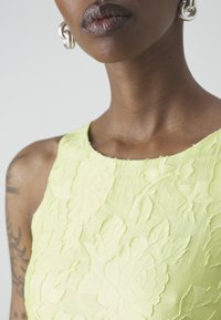 Robe sans manches vert lime avec broderies florales ; tissu texturé et encolure ronde. De grandes boucles d'oreilles en argent ajoutent du contraste.