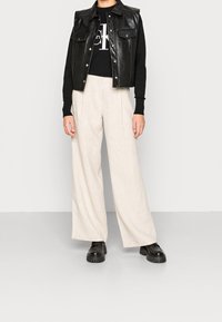 Czarna skórzana kamizelka bez rękawów na czarnym swetrze z białym logo, w połączeniu z kremowymi szerokimi spodniami i masywnymi czarnymi butami.