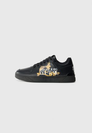 Black leather sneaker with embossed detailing, featuring gold and white text, rounded toe, and a black rubber sole. Versace Jeans Couture branding visible.