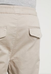 Beige cargobroek met elastische tailleband, voorzien van twee achterzakken met klep en een gladde, zachte textuur. Ontworpen voor een ontspannen pasvorm.
