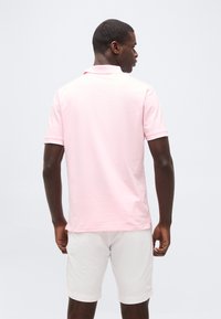 Homme de dos portant un polo à manches courtes rose clair et un short blanc sur un fond uni clair.