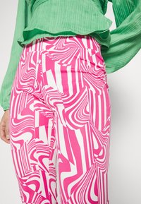 Růžové a bílé kalhoty se vzorem s odvážnými vlnitými liniemi. Materiál se zdá být elastický. Doplňuje je zelený texturovaný top.