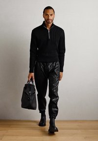 Hombre caminando por dentro, vistiendo un suéter negro con cremallera, pantalones jogger de cuero negro, botas negras y llevando una bolsa de viaje de cuero negro.