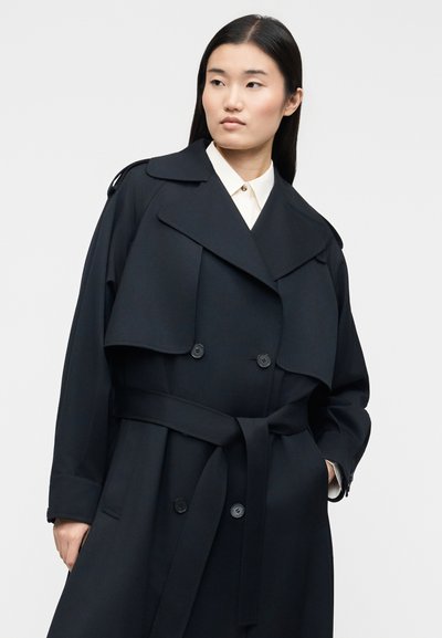 Jeune femme aux longs cheveux noirs portant un trench-coat noir ceinturé par-dessus une chemise blanche à col, regardant légèrement sur le côté.