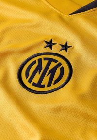 Maillot de sport jaune avec un col en V, arborant un logo rond noir et deux étoiles noires au-dessus, fabriqué en tissu lisse et texturé.